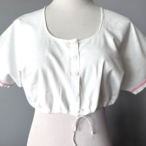 May include: White cropped top with a scoop neckline and short sleeves. The top features a button-down front and a drawstring at the waist. The sleeve edges are trimmed with a thin pink stripe. The top is displayed on a mannequin.