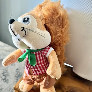 May include: A plush squirrel toy wearing a red and white checked shirt with a green bow tie and brown shorts. The squirrel has a bushy tail and a friendly face.