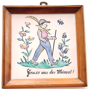 Könnte beinhalten: Eine gerahmte Fliese mit einer bunten Illustration eines Jungen, der auf einem Feld mit Blumen und Schmetterlingen spazieren geht. Die Fliese trägt den Text "Gruss aus der Heimat!" in Kursivschrift.