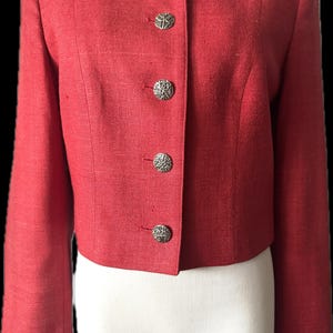 May include: A cropped, long-sleeved jacket in a vibrant red hue with a textured fabric finish. The garment features a round neckline and a front closure secured by five circular, silver-toned metal buttons with an intricate, embossed pattern.
