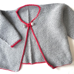 May include: A gray knitted cardigan with red trim and a red tie closure. The cardigan has short sleeves and two silver-colored buttons at the closure. The edges of the cardigan are trimmed with red yarn.