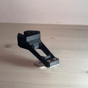 Puede incluir: Un soporte de cámara impreso en 3D de color negro con dos tornillos dorados. El soporte está diseñado para sujetarse a un trípode de cámara.