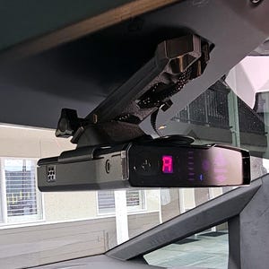 Könnte beinhalten: Ein schwarzer Radarwarner, der im Inneren eines Fahrzeugs an der Windschutzscheibe befestigt ist. Das Gerät zeigt eine rote Digitalanzeige und hat eine Reihe von Kontrollleuchten. Die Halterung ist schwarz und scheint maßgefertigt zu sein.