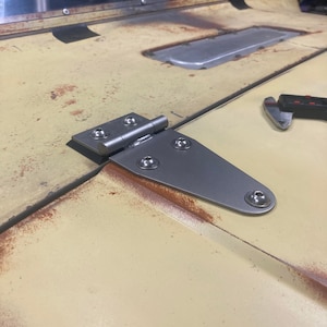 Puede incluir: Primer plano de una bisagra de metal plateado con una placa triangular y varios tornillos. La bisagra está unida a una superficie pintada de amarillo con manchas de óxido.