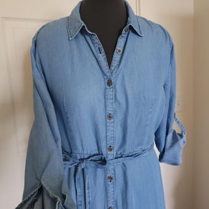 May include: Light blue denim shirt dress with long sleeves and a tie belt. The dress features a button-down front, a collared neckline, and buttoned sleeve cuffs. The dress is displayed on a black mannequin.