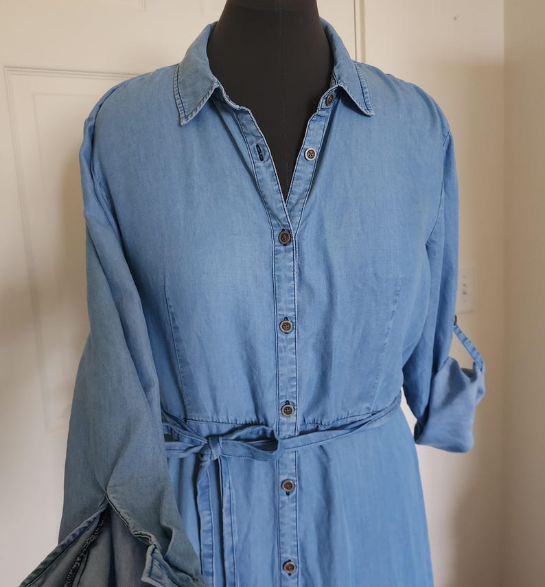 May include: Light blue denim shirt dress with long sleeves and a tie belt. The dress features a button-down front, a collared neckline, and buttoned sleeve cuffs. The dress is displayed on a black mannequin.