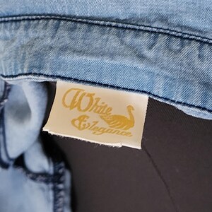 May include: Close-up of a light blue denim garment. A rectangular tag with the words "White & Elegance" and a swan graphic is attached to the garment. The tag is cream-colored with gold lettering.