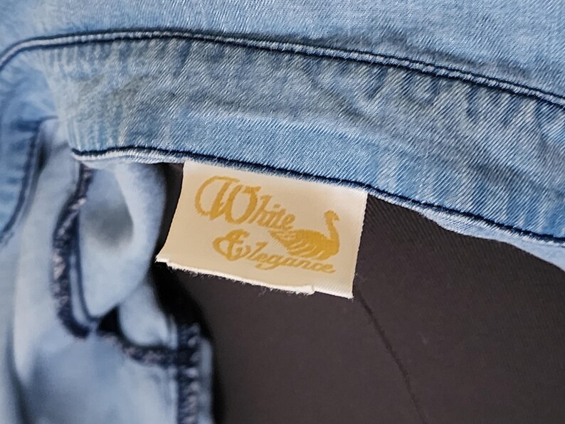 May include: Close-up of a light blue denim garment. A rectangular tag with the words "White & Elegance" and a swan graphic is attached to the garment. The tag is cream-colored with gold lettering.