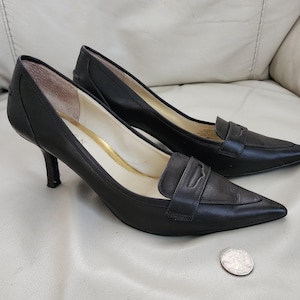 May include: A pair of black leather pumps with pointed toes and small heels. The shoes have a classic loafer-style design with a strap across the top. A silver coin is next to the shoes.