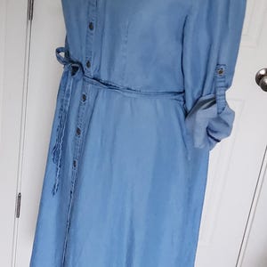 May include: Light blue denim dress with a button-down front, long sleeves, and a tie waist. The dress has a collared neckline and is displayed on a black mannequin. The sleeves are rolled up, and the dress is floor-length.
