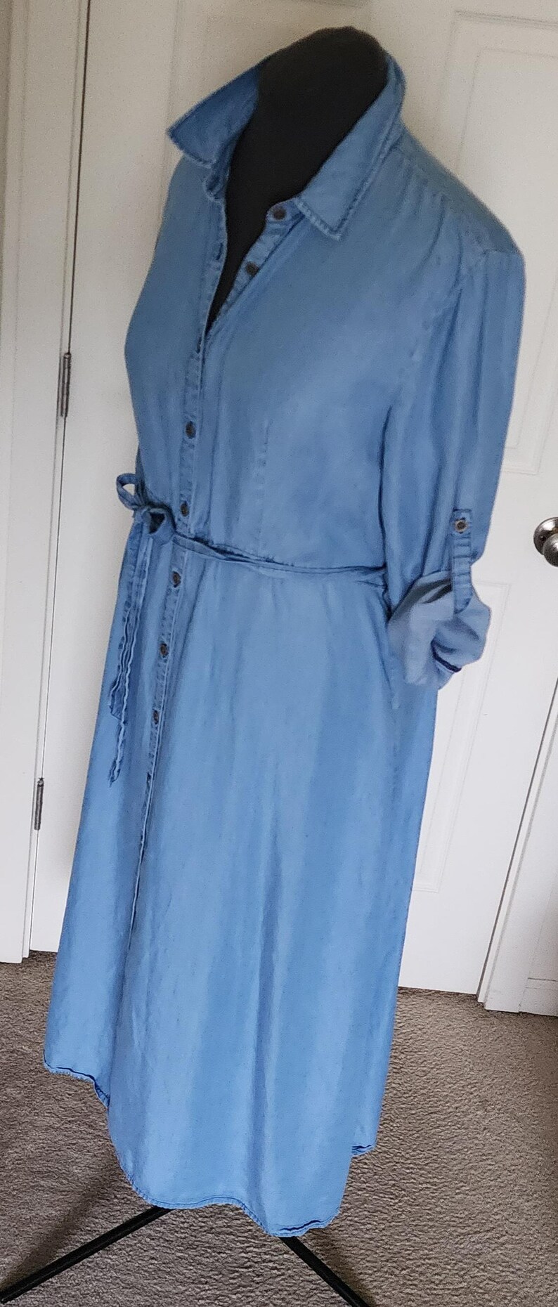 May include: Light blue denim dress with a button-down front, long sleeves, and a tie waist. The dress has a collared neckline and is displayed on a black mannequin. The sleeves are rolled up, and the dress is floor-length.