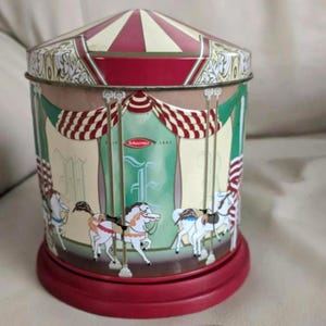 May include: A cylindrical metal tin featuring a carousel design with a red and white striped conical lid. The body displays green and cream panels, red and white draped curtains, and white horses on poles. The text Schuermer 1849 is visible.