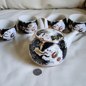 May include: A ceramic tea set consisting of a side-handled teapot and four matching cups. Each piece features a black background with white dragon illustrations, gold accents, and cloud motifs. A US quarter coin is placed for scale reference.
