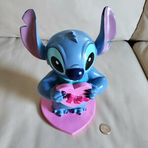 May include: A blue Stitch figurine holding a pink heart-shaped box with a pink bow. The figurine has large ears with purple tips and is mounted on a pink heart-shaped base. A coin is visible.
