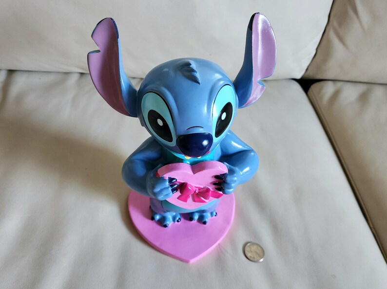 May include: A blue Stitch figurine holding a pink heart-shaped box with a pink bow. The figurine has large ears with purple tips and is mounted on a pink heart-shaped base. A coin is visible.