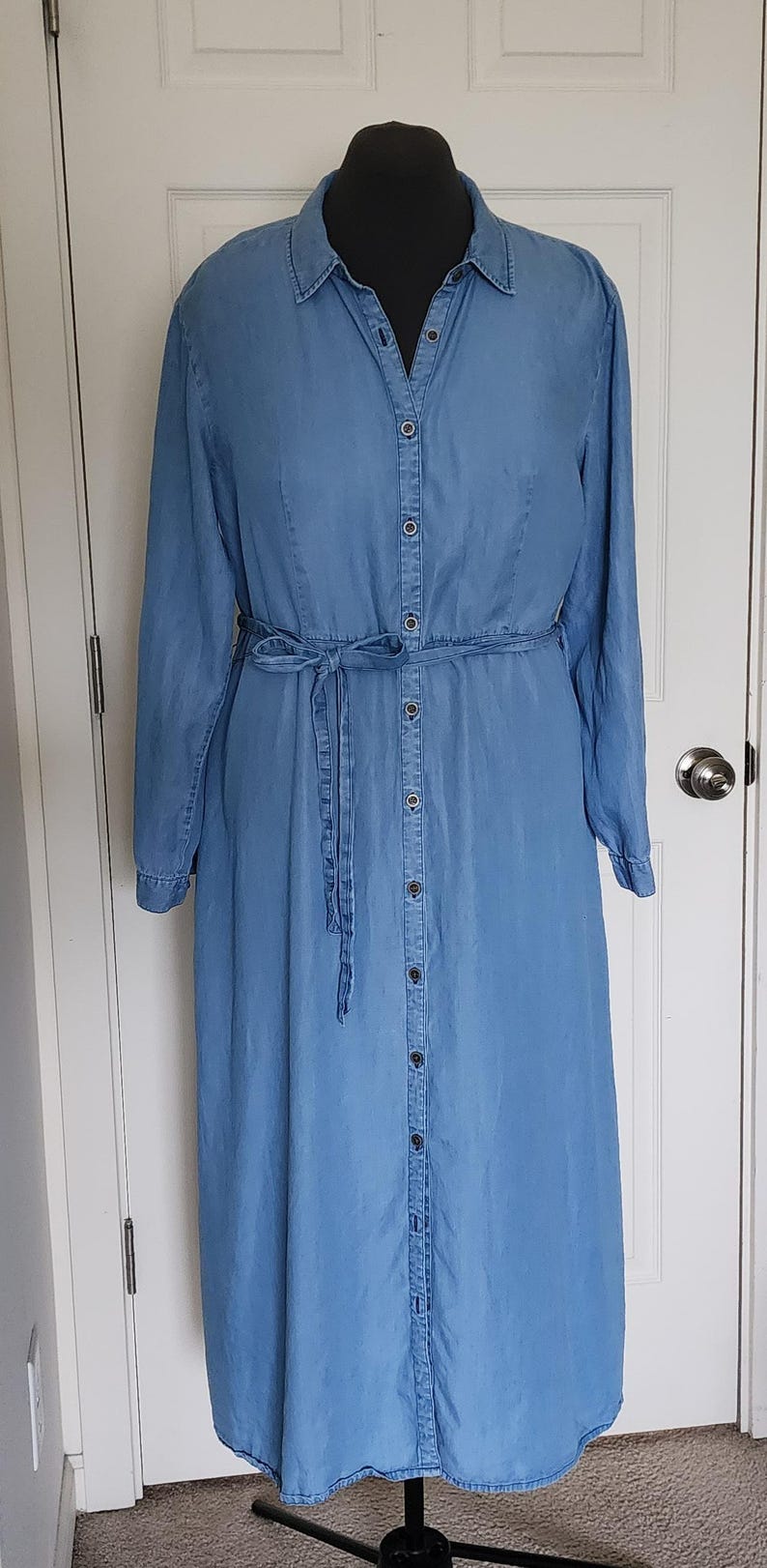 May include: A long, light blue denim dress with long sleeves and a button-down front. The dress features a matching fabric belt tied at the waist. The dress is displayed on a black mannequin against a white door.