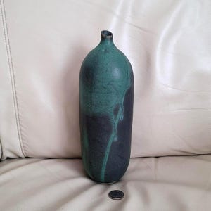 May include: A tall, ceramic vase with a narrow neck and rounded body. The vase is primarily dark teal, with a darker, almost black, base. Teal streaks run down the side, creating a unique pattern. A coin rests near the base.