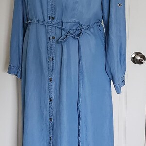 May include: A light blue denim dress with long sleeves, a button-down front, and a tie waist. The dress is displayed on a black mannequin and has a midi length. The sleeves have buttoned cuffs. The dress is made of a soft denim material.