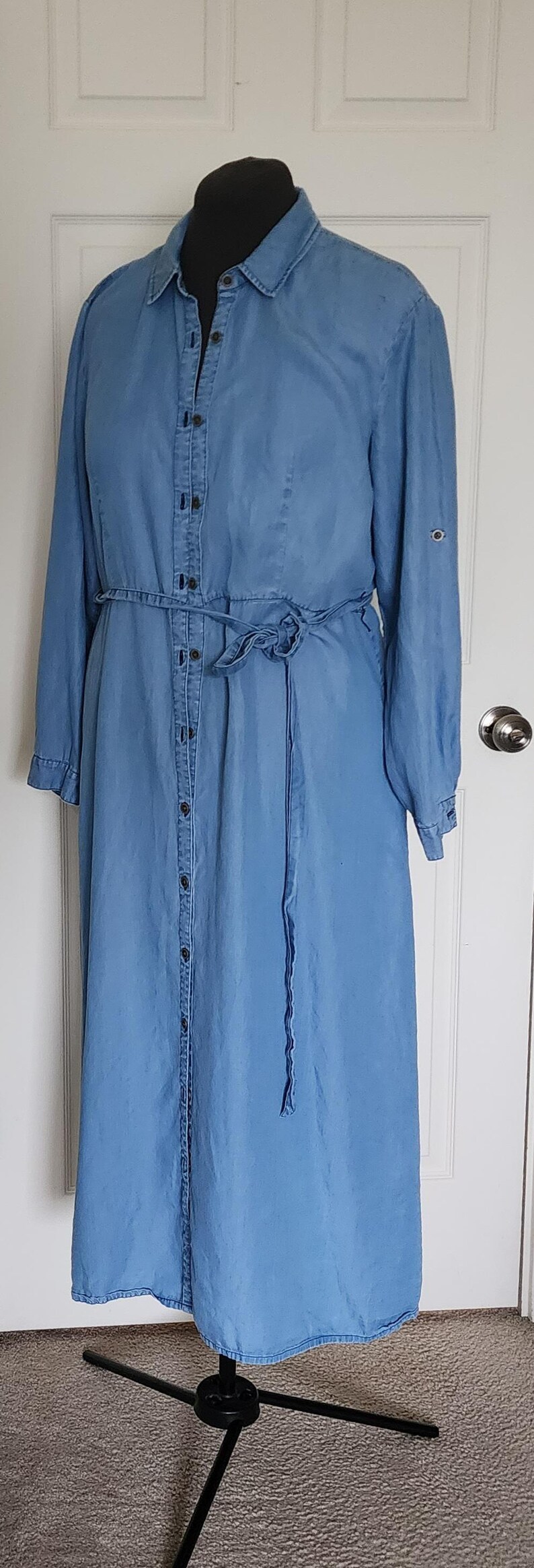 May include: A light blue denim dress with long sleeves, a button-down front, and a tie waist. The dress is displayed on a black mannequin and has a midi length. The sleeves have buttoned cuffs. The dress is made of a soft denim material.