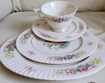 May include: A white porcelain tea set consisting of a teacup, a saucer, and two plates of varying sizes. Each piece features a delicate fluted edge and a colorful floral pattern with pink, yellow, and blue flowers and green leaves.