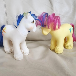 Puede incluir: Dos figuras de poni de plástico. El poni de la izquierda es blanco con crin azul y amarilla, con un símbolo de escalera roja en el flanco. El poni de la derecha es amarillo con crin rosa y morada, con un símbolo de cactus azul.