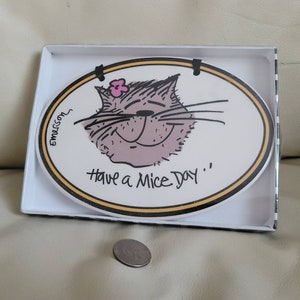 May include: An oval ceramic plaque featuring a cartoon cat illustration with a pink flower, the text "Have a Mice Day", and the signature "Emerson". The plaque has a black and yellow border and is displayed in a white box with a US coin for scale.