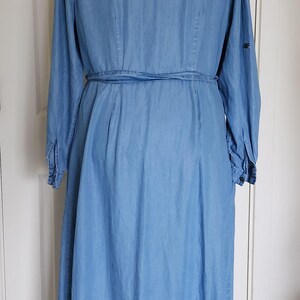 May include: Light blue denim dress with long sleeves and a matching belt. The dress has a button-down front, a collared neckline, and a flared skirt. The dress is displayed on a black dress form.