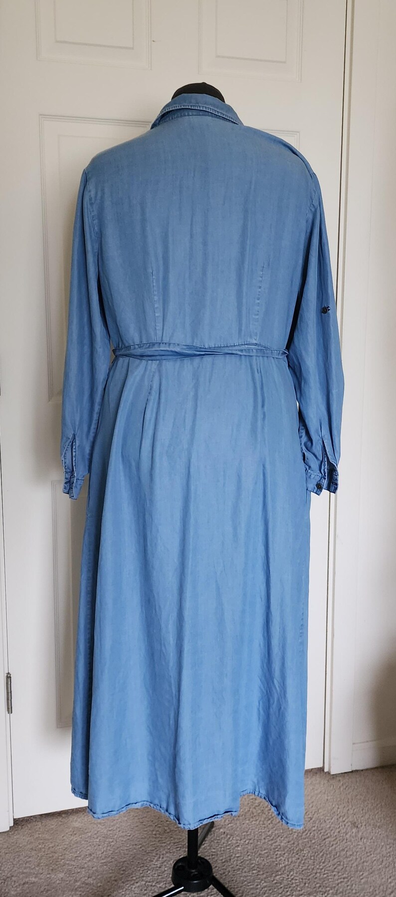 May include: Light blue denim dress with long sleeves and a matching belt. The dress has a button-down front, a collared neckline, and a flared skirt. The dress is displayed on a black dress form.