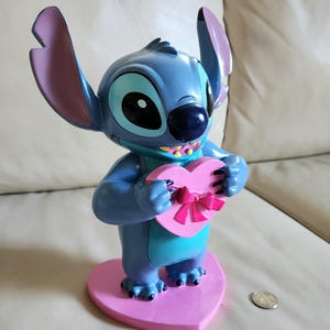 May include: A blue Stitch figurine holding a pink heart-shaped box with a pink bow. The figurine has large ears with purple tips and is standing on a pink heart-shaped base. The figurine is approximately 6 inches tall.