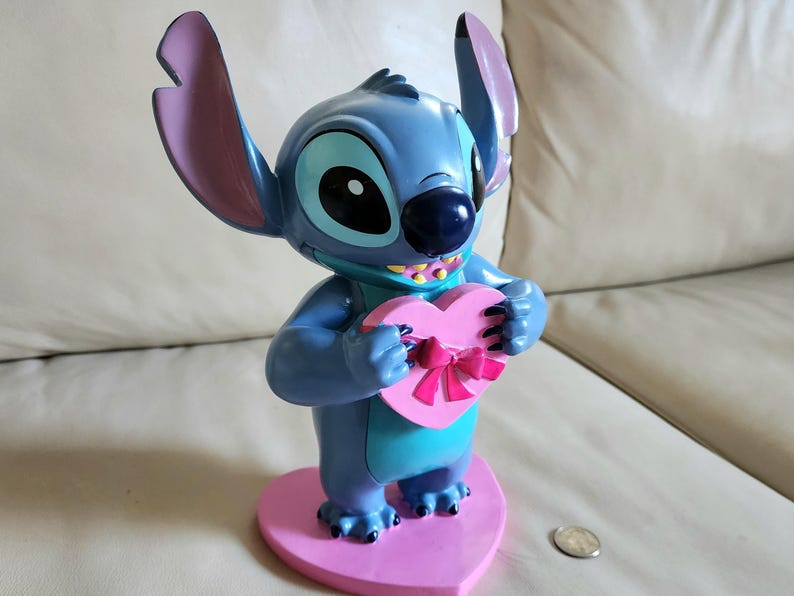 May include: A blue Stitch figurine holding a pink heart-shaped box with a pink bow. The figurine has large ears with purple tips and is standing on a pink heart-shaped base. The figurine is approximately 6 inches tall.