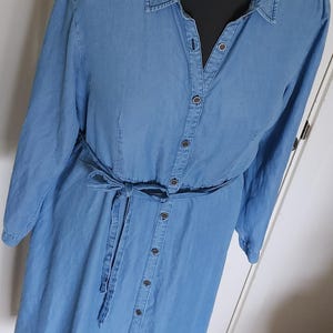 May include: A light blue denim dress with long sleeves and a button-down front. The dress features a matching fabric belt tied at the waist. The dress is displayed on a black mannequin.
