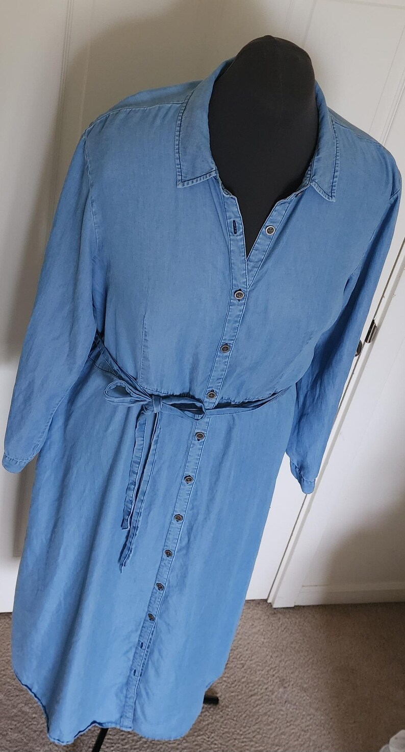 May include: A light blue denim dress with long sleeves and a button-down front. The dress features a matching fabric belt tied at the waist. The dress is displayed on a black mannequin.
