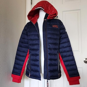 May include: A navy blue puffer jacket with a red hood and red accents on the sleeves. The jacket has a full-length zipper and the Tommy Hilfiger logo embroidered on the chest. The jacket is displayed on a white mannequin.