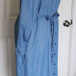 May include: A light blue denim shirt dress with a button-down front, long sleeves, and a tie belt. The dress is displayed on a black mannequin and has a collar and pockets.