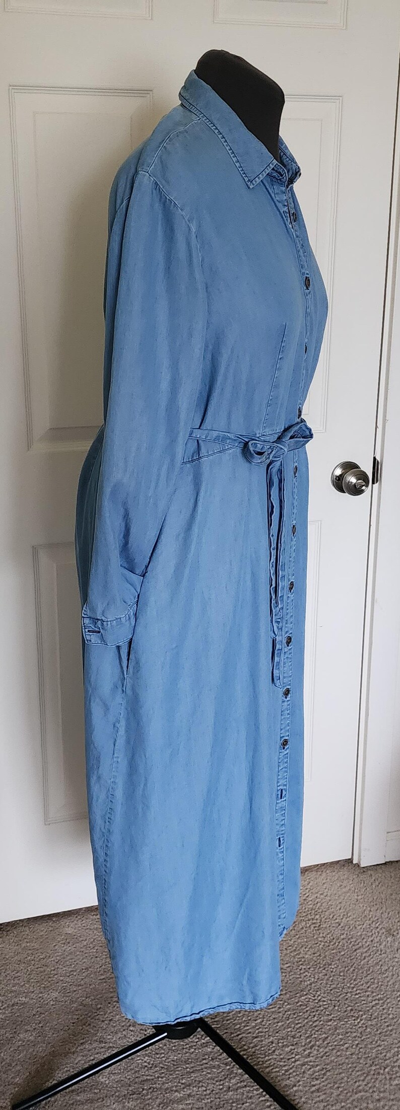 May include: A light blue denim shirt dress with a button-down front, long sleeves, and a tie belt. The dress is displayed on a black mannequin and has a collar and pockets.