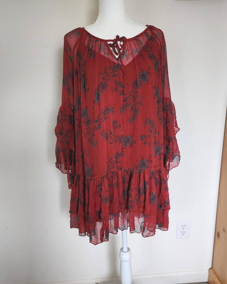 May include: A red dress with a floral design. The dress features long, flowing sleeves and a tiered, ruffled hem. The neckline is a scoop with a tie detail. The fabric is sheer and lightweight, ideal for warmer weather.