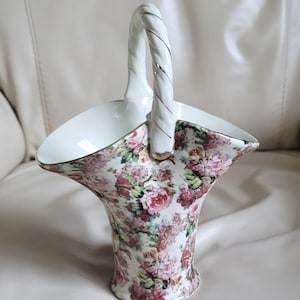 May include: A white porcelain basket featuring a vibrant floral pattern with pink, red, and green roses. The basket has a flared, scalloped rim, a twisted handle with gold trim, and a smooth white interior. The exterior is covered in flowers.