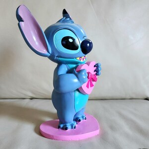 May include: A blue figurine of the character Stitch from Lilo & Stitch, holding a pink heart-shaped box with a bow. The figurine stands on a pink heart-shaped base. The figure has large ears and a friendly expression.