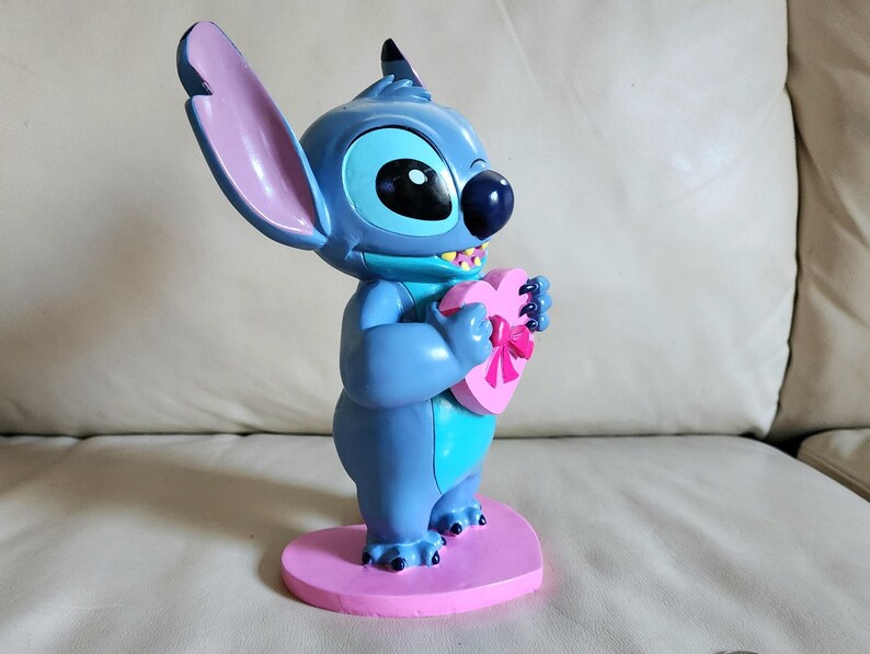 May include: A blue figurine of the character Stitch from Lilo & Stitch, holding a pink heart-shaped box with a bow. The figurine stands on a pink heart-shaped base. The figure has large ears and a friendly expression.