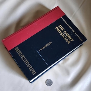 May include: A vintage hardcover book titled "The Family Physician" with a red spine and a dark blue cover. The book features gold lettering and a medical symbol. A silver coin is visible near the book.