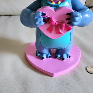 May include: A blue and teal figurine of a cartoon character holding a pink heart-shaped box with a pink bow. The figurine is on a pink heart-shaped base. A US quarter is next to the base.