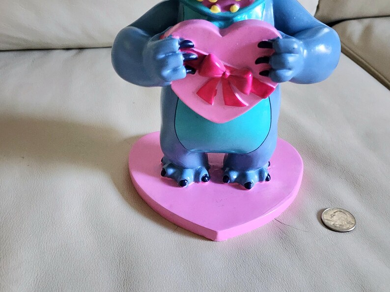 May include: A blue and teal figurine of a cartoon character holding a pink heart-shaped box with a pink bow. The figurine is on a pink heart-shaped base. A US quarter is next to the base.