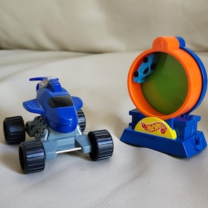 May include: Two Hot Wheels toy vehicles. One is a blue and gray car with large black wheels. The other is a blue and orange toy with a circular window and a small car inside. The Hot Wheels logo is visible.