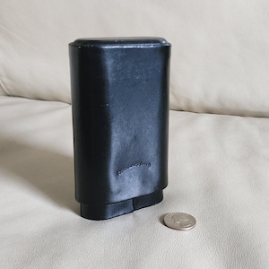 May include: A black leather cigar case with a rectangular shape and a rounded top. The case has a small, embossed logo and a two-part base. A quarter is next to the case for size comparison.