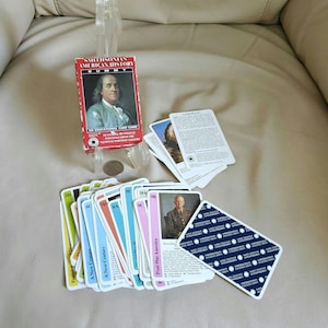 May include: A Smithsonian American History Rummy card game set featuring a red box with a portrait of a historical figure. The set includes a deck of cards with colourful borders, historical portraits, descriptive text, and a blue patterned back.