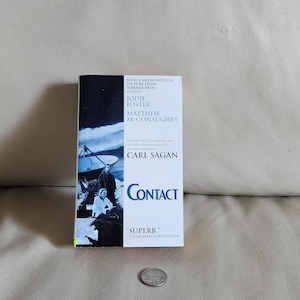 May include: A paperback book titled "Contact" by Carl Sagan. The cover features a dark blue background with a depiction of a radio telescope and two figures. The book stars Jodie Foster and Matthew McConaughey.