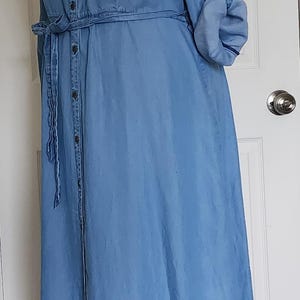 May include: A light blue denim dress with a button-down front, long sleeves, and a tie waist. The dress has a collared neckline and is displayed on a mannequin. The sleeves are rolled up, and the dress falls to the ankles.