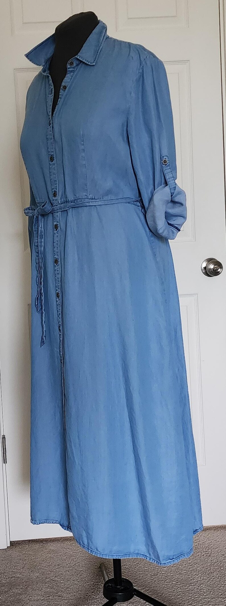 May include: A light blue denim dress with a button-down front, long sleeves, and a tie waist. The dress has a collared neckline and is displayed on a mannequin. The sleeves are rolled up, and the dress falls to the ankles.