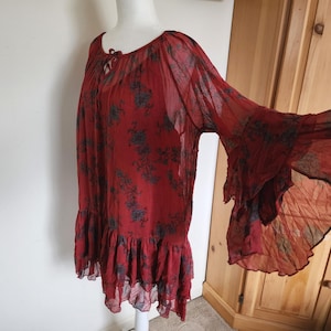 May include: A red tunic top with a floral design and bell sleeves. The top has a ruffled hem and a tie at the neckline. The fabric is sheer and lightweight. The top is suitable for warmer weather.