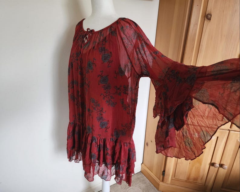 May include: A red tunic top with a floral design and bell sleeves. The top has a ruffled hem and a tie at the neckline. The fabric is sheer and lightweight. The top is suitable for warmer weather.